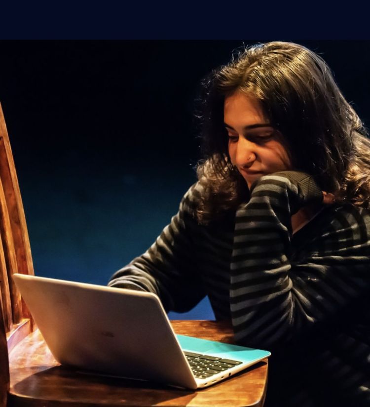 Actor is sat on the floor looking at a laptop which is balanced on a chair. The lighting is from above creating shadows.